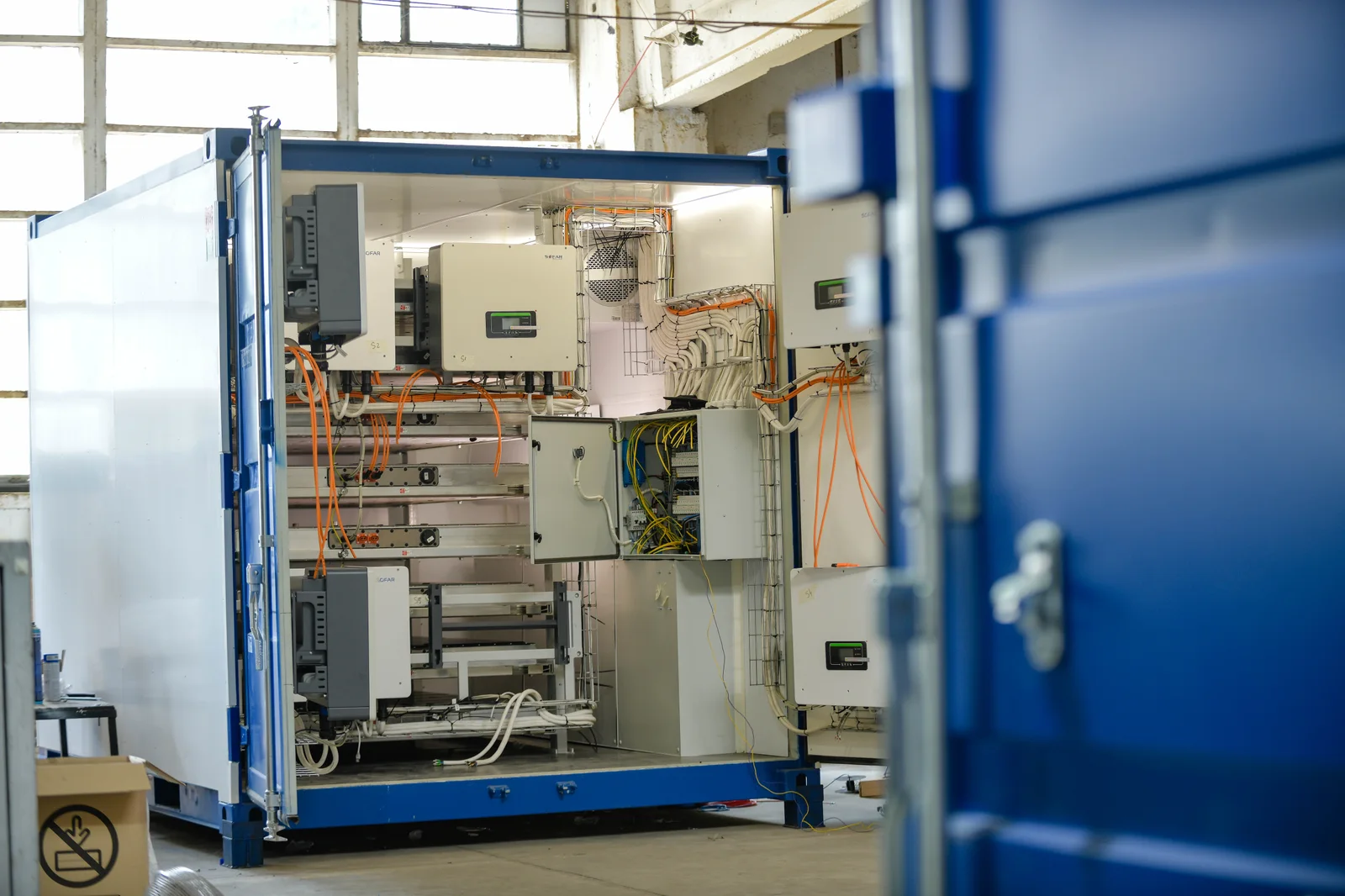 A Marny Energy Battery-Shelter in the workshop with its doors open: blue ten-foot container, hybrid inverters, battery racks and high-voltage cabling